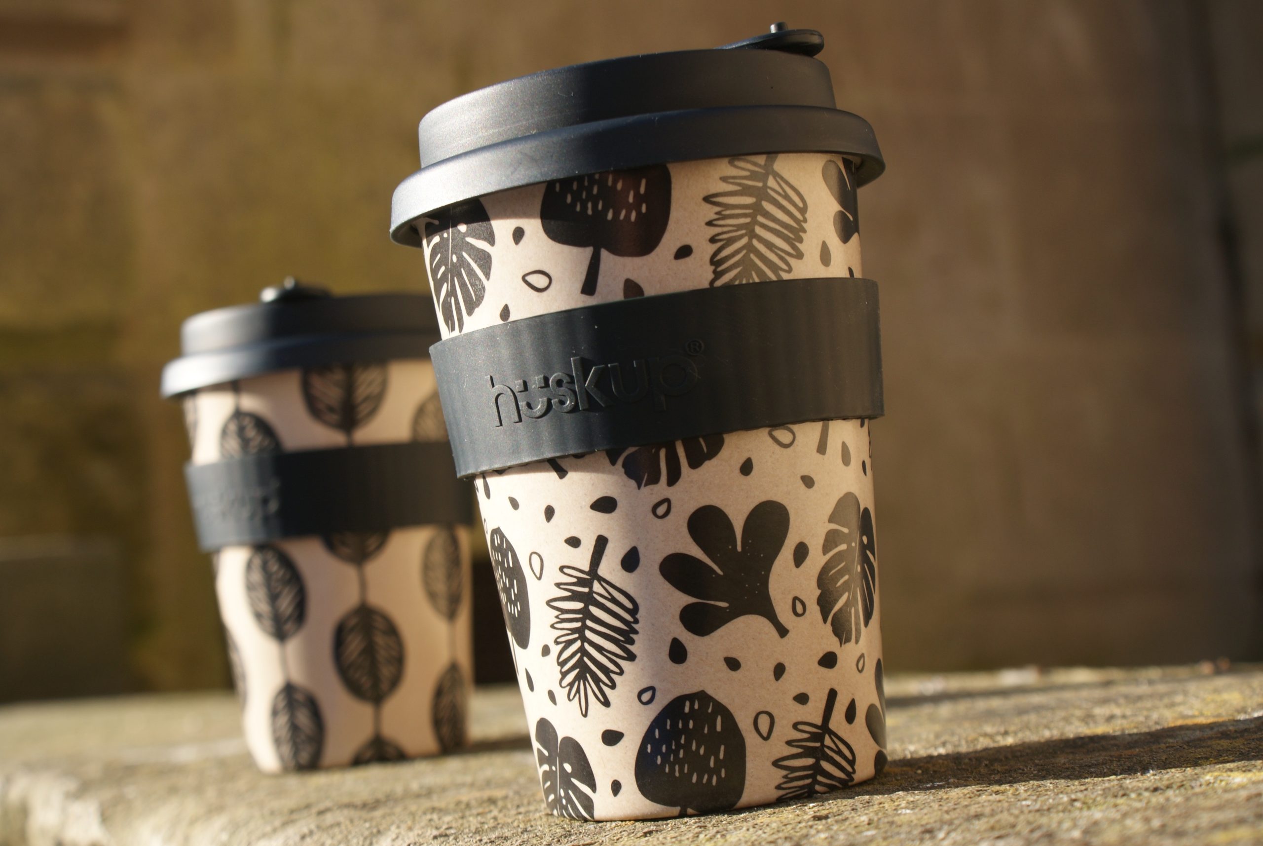 Two reusable cup huskups in the sun with black bands the one to the left has a black and brown leaf design and the one to the right has leaves and dots in different shapes and sizes the one on the right is placed closer to the camera
